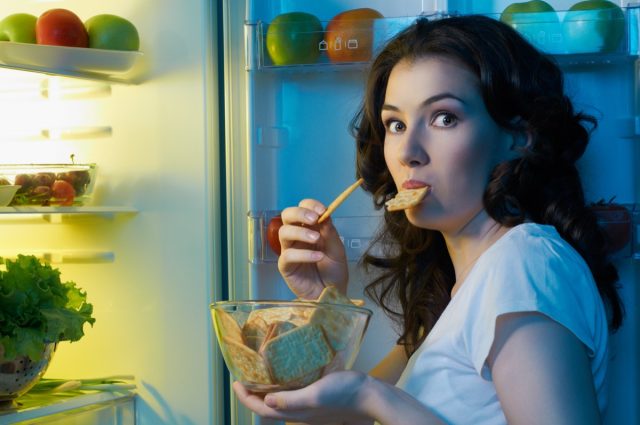 Woman eating snacks in night next to the opened fridge.
