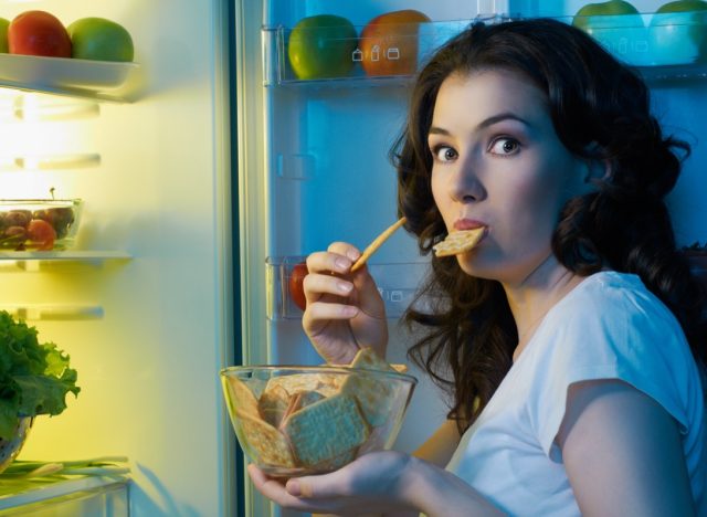 Woman eating snacks in night next to the opened fridge.