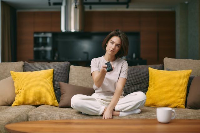 Tired bored young woman changing tv channels by remote controller, sitting on sofa at home.