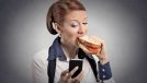 Closeup portrait young serious corporate business woman deal maker reading news message on smart mobile phone holding eating sandwich isolated grey background.