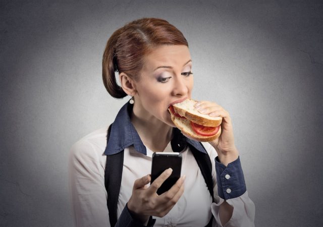 Closeup portrait young serious corporate business woman deal maker reading news message on smart mobile phone holding eating sandwich isolated grey background.