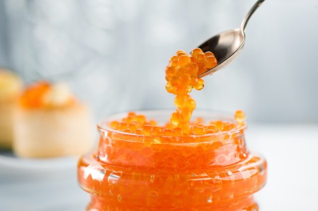 Red Caviar in a spoon, fish roe in a glass jar. Close-up of salmon fish roe caviar on served table.