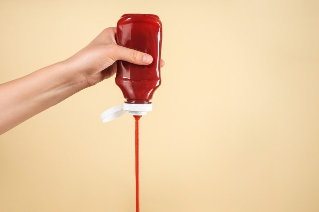 Woman pouring ketchup from bottle on beige background, closeup