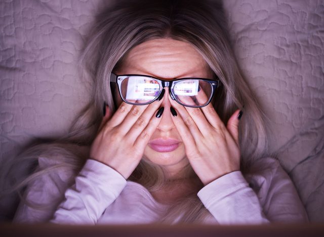 Sleepy young caucasian woman in glasses rubbing her eyes, feels tired after working on a laptop, lying on pillow, front view/ Overwork, tired, health concept/ Exhausted and fatigue eyes