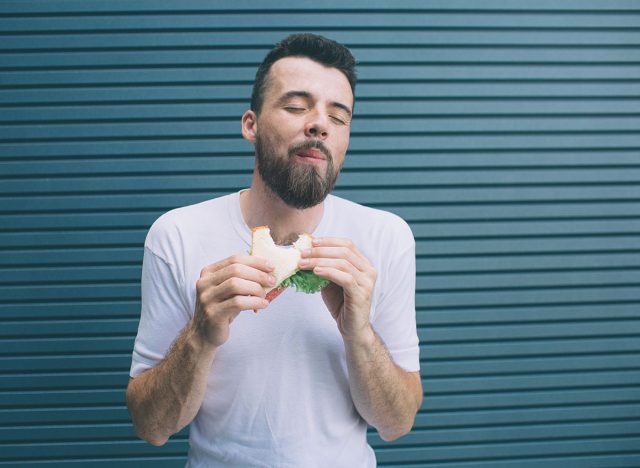 A picture of delightful man enjoying his meal. He is chewing a piece of sandwich and keeping eyes closed. Isolated on striped and blue background.