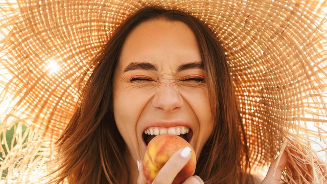 Photo of young positive happy cheery woman in hat posing outdoors at the nature green grass near beach eat peach.