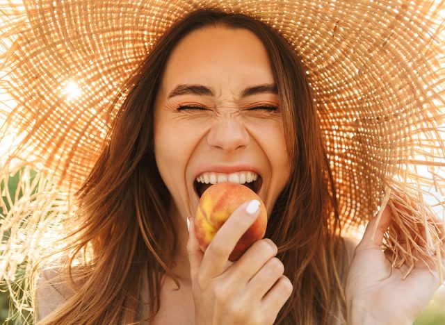 Photo of young positive happy cheery woman in hat posing outdoors at the nature green grass near beach eat peach.