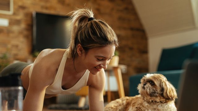 Young happy athletic woman exercising in plank position while a dog is relaxing next to her in the living room.