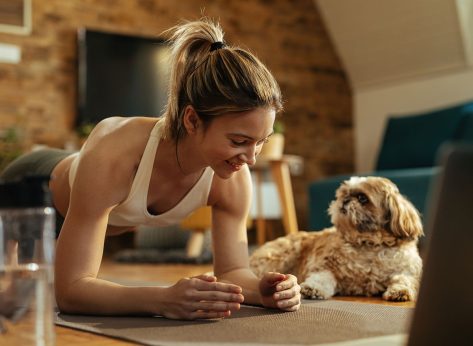 Young happy athletic woman exercising in plank position while a dog is relaxing next to her in the living room.
