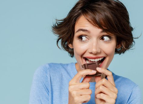 Close up young happy woman in casual sweater hold sweet pink cream donuts biting chocolate bar look camera isolated on plain pastel light blue background studio portrait. People lifestyle food concept