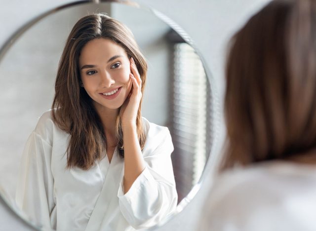 Self-Care Concept. Young Attractive Woman Looking At Mirror In Bathroom, Pretty Female Touching Face And Smiling To Reflection, Enjoying Her Beautiful Smooth Skin, Selective Focus, Closeup