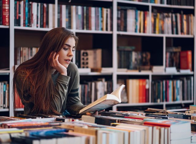 Beautiful young woman buying books at a bookstore and reading one. Geeky woman reading a book with a bookshelves in background.