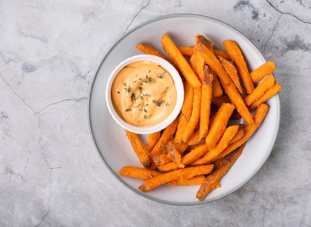 Baked sweet potato fries on a plate with savory sauce over concrete background, top view