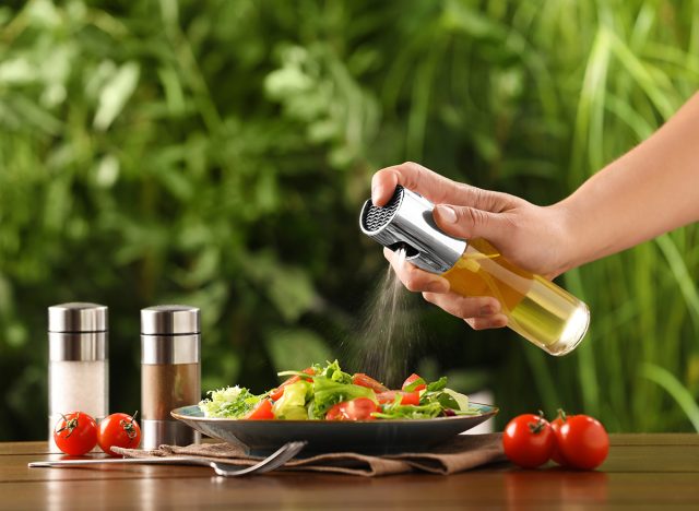 Woman spraying cooking oil onto delicious salad at wooden table against blurred green background, closeup