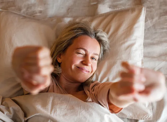 Good morning, new day, weekend, holiday. Happy middle aged woman sits on bed, lady stretching arms after sleep and enjoying morning in cozy comfort bedroom interior, free space