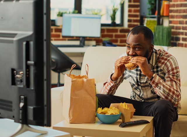 African american guy having fun eating burgers and drinking beer, watching comedy movie on television. Male adult enjoying fast food delivery and alcohol, watch tv show in living room.