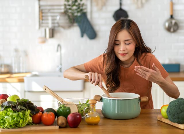 Young woman standing near stove and cooking, housewife, meal, chef, food.Happy woman looking and smelling tasting fresh delicious from soup in a pot with steam at white interior kitchen