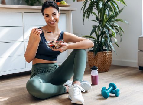 Shot of athletic woman eating a healthy bowl of muesli with fruit sitting on floor in the kitchen at home