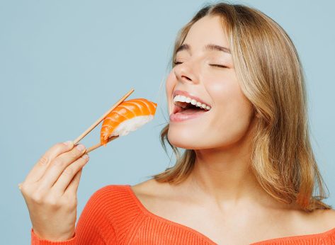 Close up young woman wear orange casual clothes close eyes open mouth hold eat raw fresh sushi roll Japanese food with chopsticks isolated on plain blue background studio portrait. Lifestyle concept