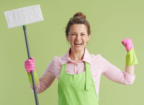 Spring cleaning. happy modern 40 years old housewife in green apron and rubber gloves against green background with mop with raised arms rejoicing.