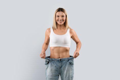 Happy cheerful attractive blonde middle aged woman wearing big jeans after weight loss on grey background