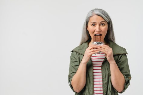 Mature middle-aged woman with grey hair eating chocolate bar.