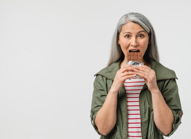 Mature middle-aged woman with grey hair eating chocolate bar.