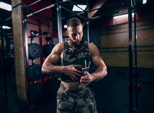 Muscular caucasian bearded man tightening up military style weighted vest in gym. Weight plates and kettlebells in background.