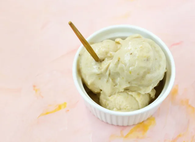 Yellow vegan homemade banana and mango dairy-free gelato ice cream in a porcelain bowl with a spoon, pink background. Copy space, selective focus.