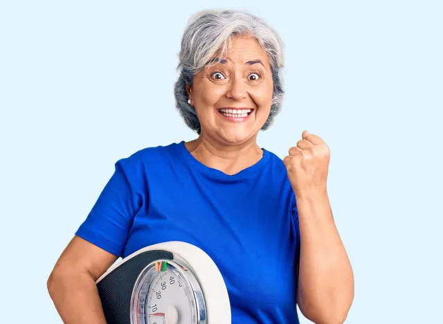 Senior woman with gray hair holding weight machine to balance weight loss screaming proud, celebrating victory and success very excited with raised arms