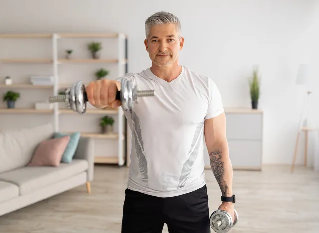 Domestic training with weights. Positive mature man doing exercises with dumbbells, strengthening his body at home. Senior sportsman working on his biceps muscles, staying healthy