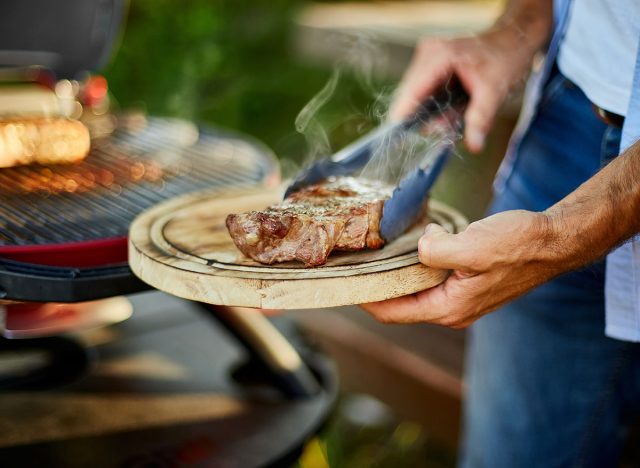 Man put down on a wooden board ready to eat grilled steak meat, male roasted steak meat on the gas grill on barbecue grill outdoor in the backyard, summer family picnic, food on the nature.