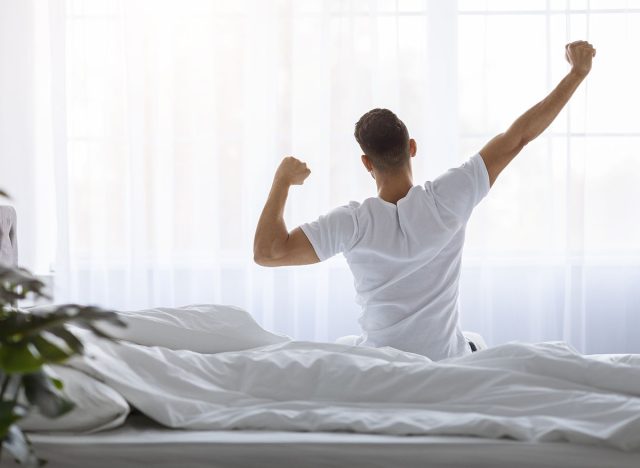 Rear View Of Young Man Stretching In Bed After Waking Up In The Morning, Unrecognizable Male Resting In Light Bedroom After Good Sleep, Looking At Window, Enjoying Start Of New Day, Copy Space