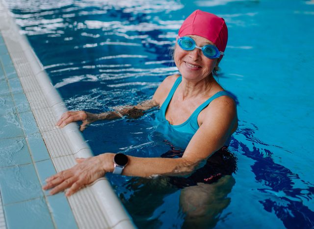 Happy senior woman in swimming pool, leaning on edge and looking at camera.