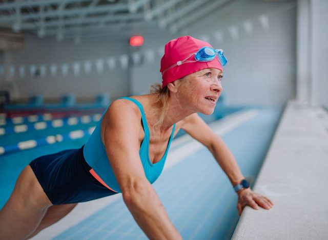 Active senior woman doing warming up exercise indoors in public swimming pool.