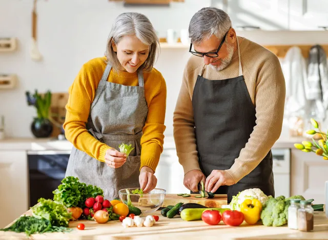 Happy elderly couple smiling husband and wife in aprons prepare salad together at kitchen table, chopping variety of colorful vegetables, trying to maintain healthy lifestyle eating vegetarian food