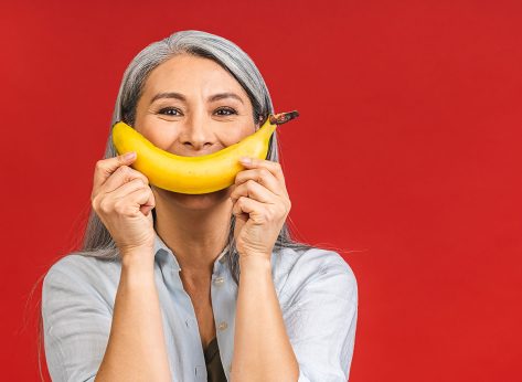 Vegan or vegetarian concept. Portrait of a beautiful elderly asian mature aged woman holding banana fruit, smiling, isolated over red background.