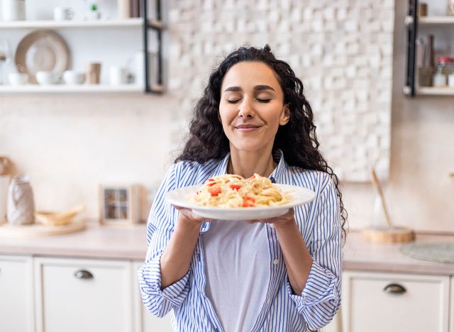 Happy lady tasting homemade spaghetti while having lunch, enjoying the smell with eyes closed, sitting in kitchen interior. Woman cooking and eating tasty food at home