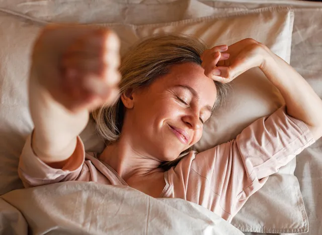 Good morning, new day, weekend, holiday. Happy middle aged woman lying on bed, lady stretching arms after sleep and enjoying morning in cozy comfort bedroom interior, free space