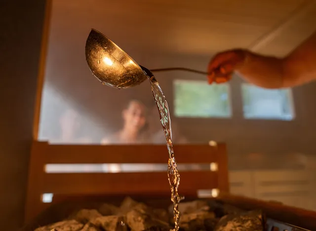 Man pouring water onto hot stone with metal spo in sauna room with a group of people. Steam an water on the stones, spa and wellness concept, relax in hot finnish sauna. Warm temperature bath therapy.