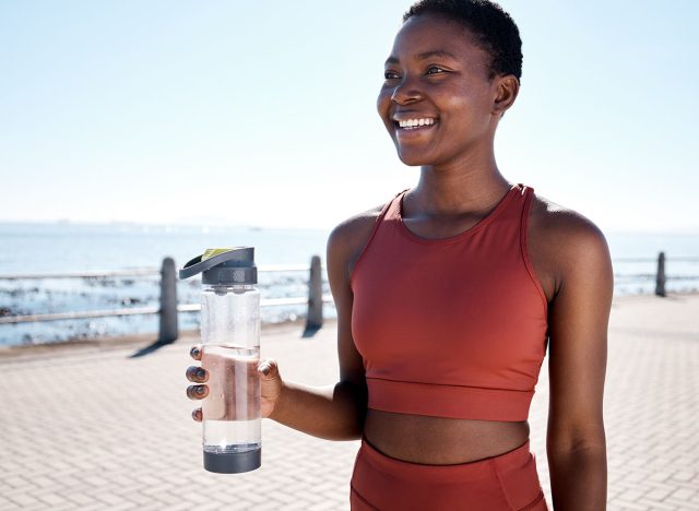 Fitness, water and black woman runner at beach for training, wellness and cardio by ocean, smile and happy. Exercise, drinking water and woman relax after running, workout and walk with hydration