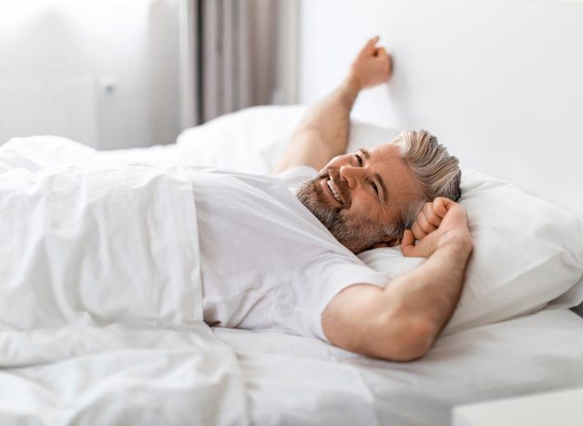 Positive cheerful handsome grey-haired bearded european mature man in white t-shirt stretching in bed in the morning, looking at copy space and smiling, feeling powerful, side view, home interior