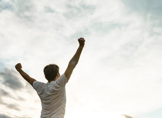 Young man standing with his arms raised high in triumph under white cloudy sky with plenty of copy space.