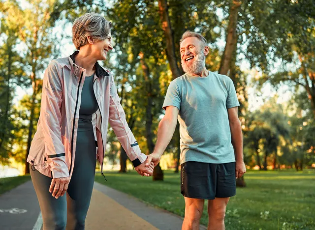 Cheerful modern senior couple outdoors in the park dressed in sportswear walking holding hands and laughing. Morning sun rays fall on smiling man's face.