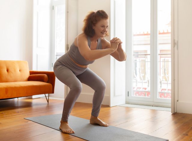 Smiling senior woman making squat exercise at home, active sporty elderly lady training on her yoga mat, smiling during fitness workout in her airy, well-lit living room, copy space