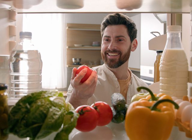 Point of view from inside refrigerator POV Caucasian man householder homeowner open fridge door kitchen full of products food smiling find take red tomato happy smile cooking salad healthy vegetarian