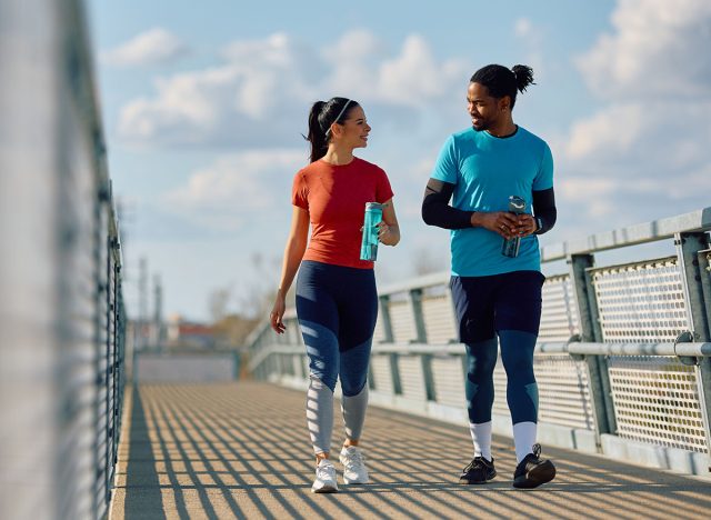 Happy athlete and her African American friend drinking water and communicating while walking over the bridge outdoors.