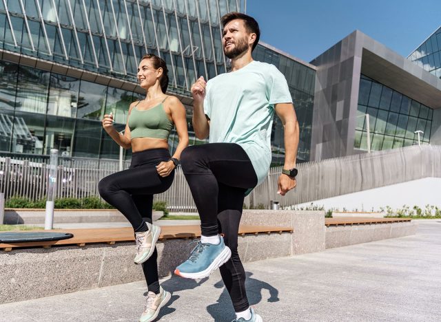 Fit couple doing high-knee exercises in front of a modern building on a clear day.