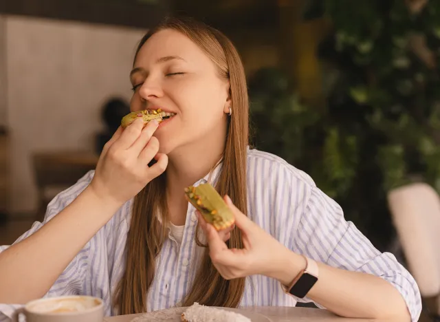 Young blonde woman eating eclair sitting in cafe. Girl bite piece of croissant look joyful at restaurant. Cheat meal day concept. Woman is preparing with appetite to eat eclair. Enjoy pistachio bakery