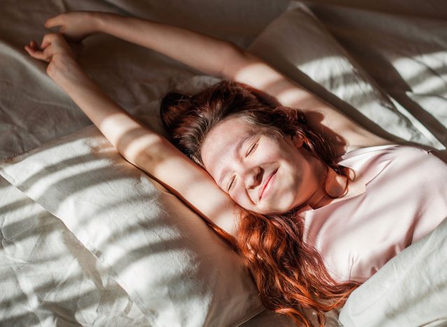 Good morning, new day, weekend, holiday. Happy Young Woman sets on Bed, teen Girl Stretching arms after sleep and enjoying morning in cozy comfort bedroom interior